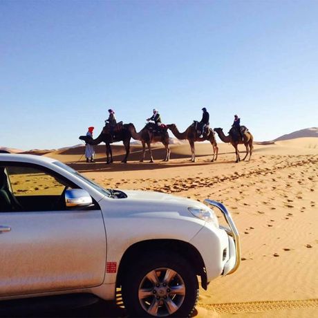 Blick auf einen Gelaendewagen vor Personen auf Kamelen in der Wueste Marokkos