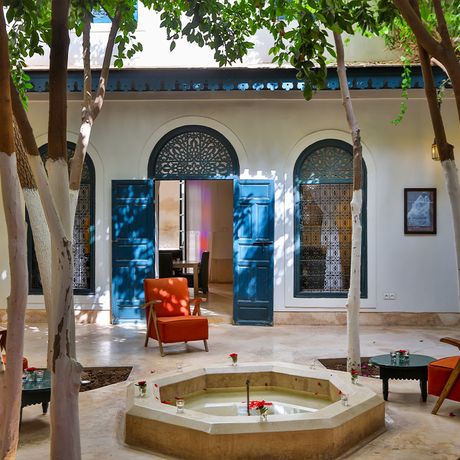 Blick auf einen Brunne in der Mitte eines Sitzbereiches zwischen Baeumen im Innenhof des Riad Dar Sara