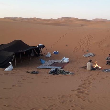 Blick auf sitzende Personen an einem Lagerfeuer vor einem Berberzelt in der Wueste