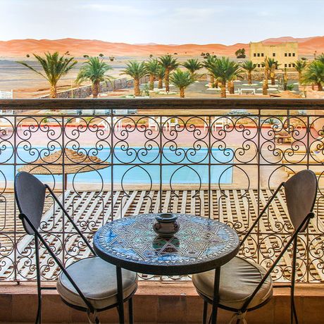 Blick auf den Balkon mit zwei Stuehlen, einem Tisch und dem Blick ueber die Duenen von Merzouga in einer Suite